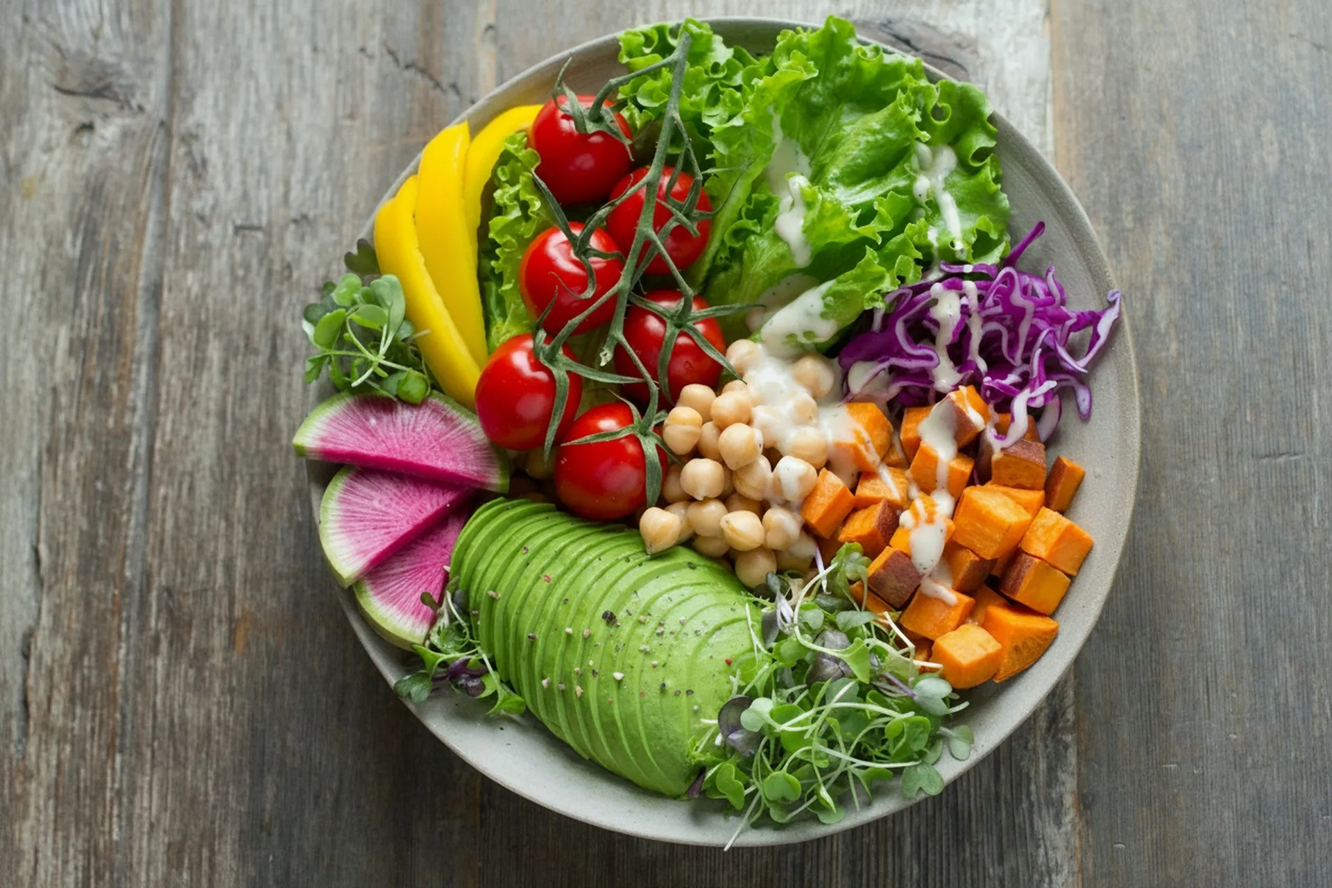 Bowl mit Avocado, Kirschtomaten, Kichererbsen, Süßkartoffel und Salat auf einem Holztisch