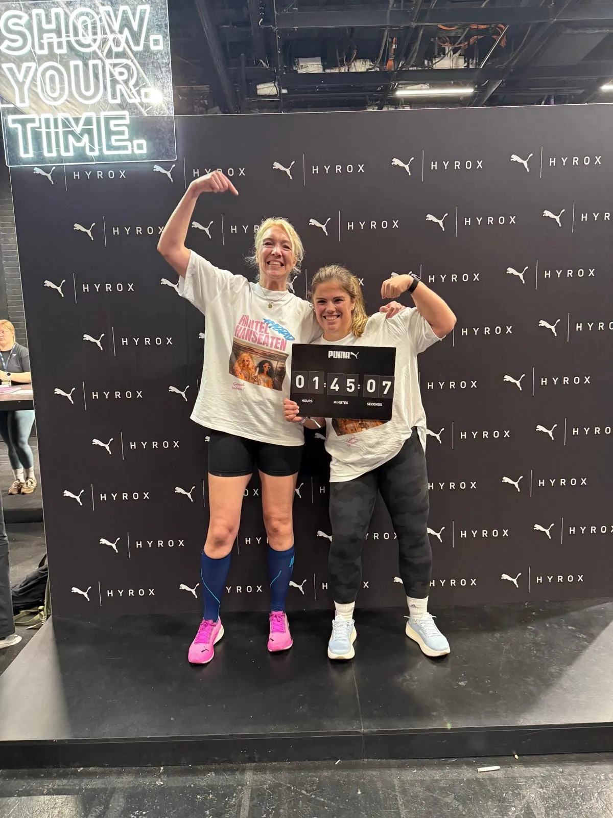 Zwei Athletinnen vor der Hyrox-Wand mit ihrer Zielzeit von 1:45:07