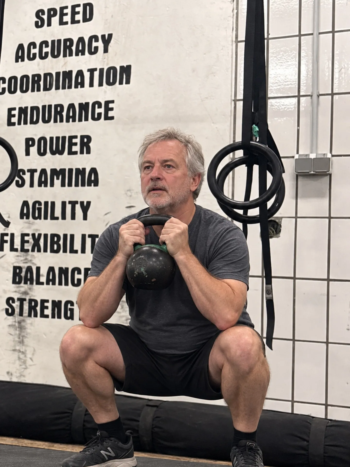 Mann Mitte 50 bei einer Kettlebell-Kniebeuge (Goblet Squat) vor Skills-Wand und Ringen